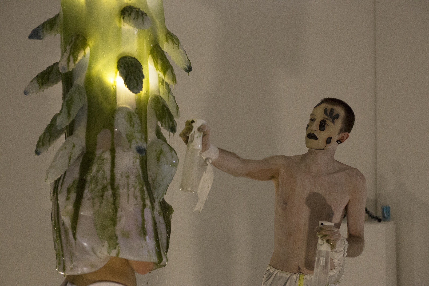 A man engulfed in a gelatenous form covered in tentacles knealing over a pool of green slime while a shirtless man painted in white sprays him with moisture